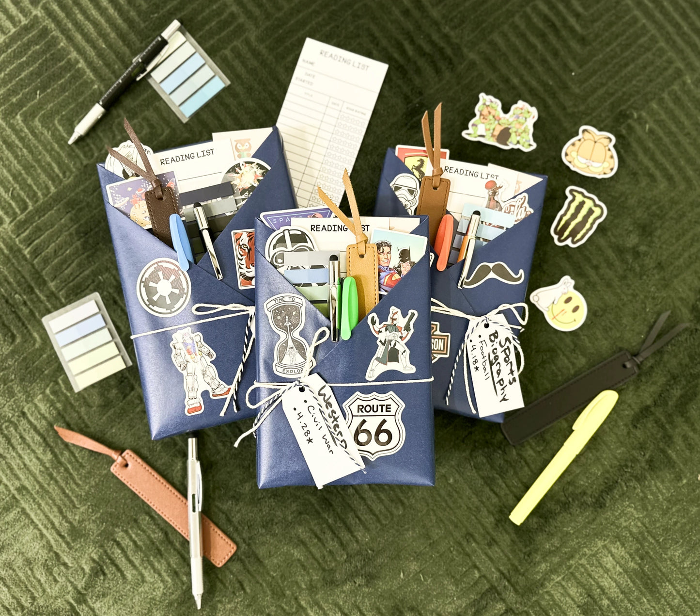 Three Blind Date with Books laid out on a dark green patterned fabric. Each book is wrapped in blue craft paper and includes different reading accessories related to the book's genre and geared toward men. Displayed accessories surround the 3 books.